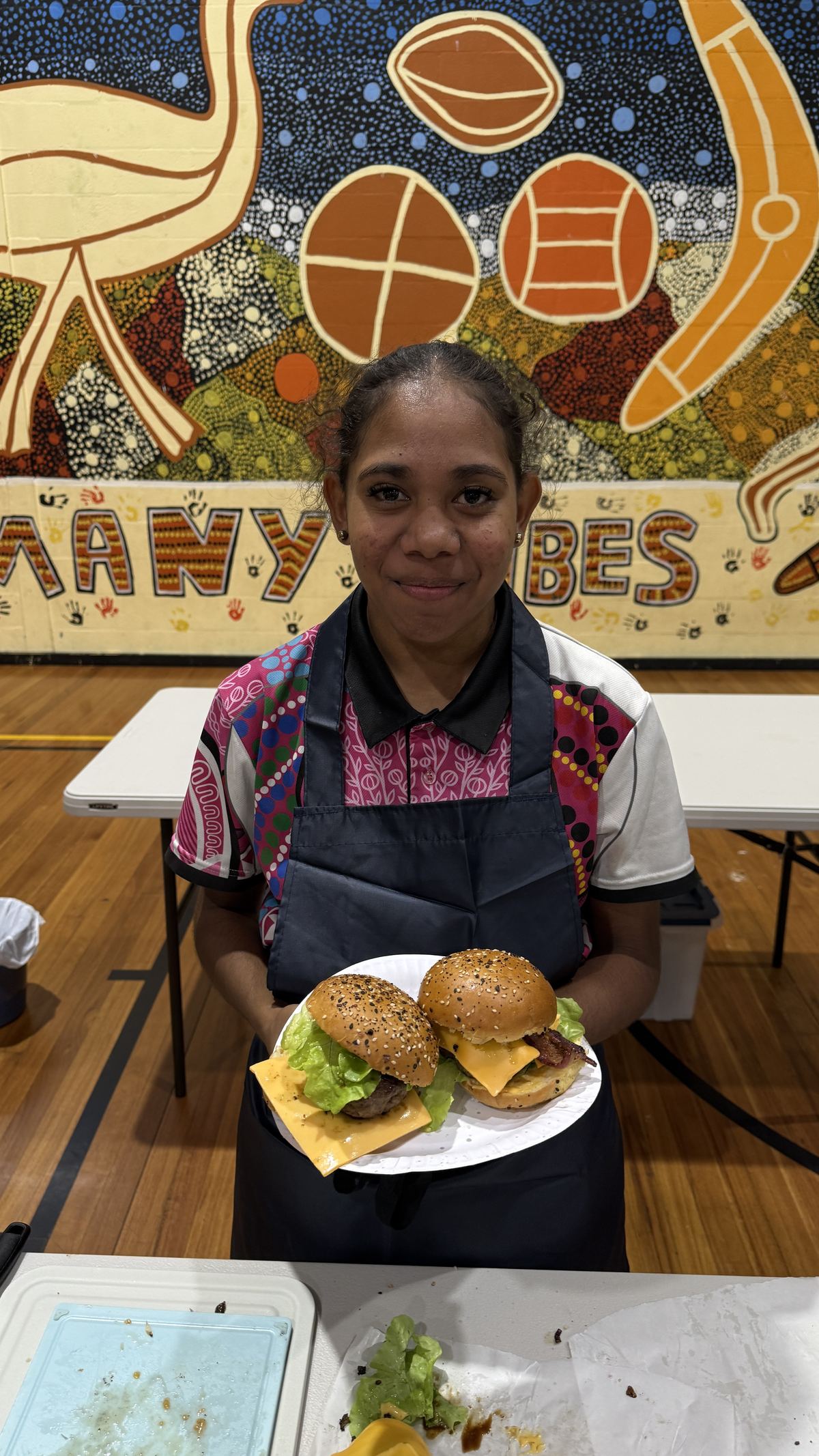 A young cook holding a plate of burgers in front of a Many Tribes mural
