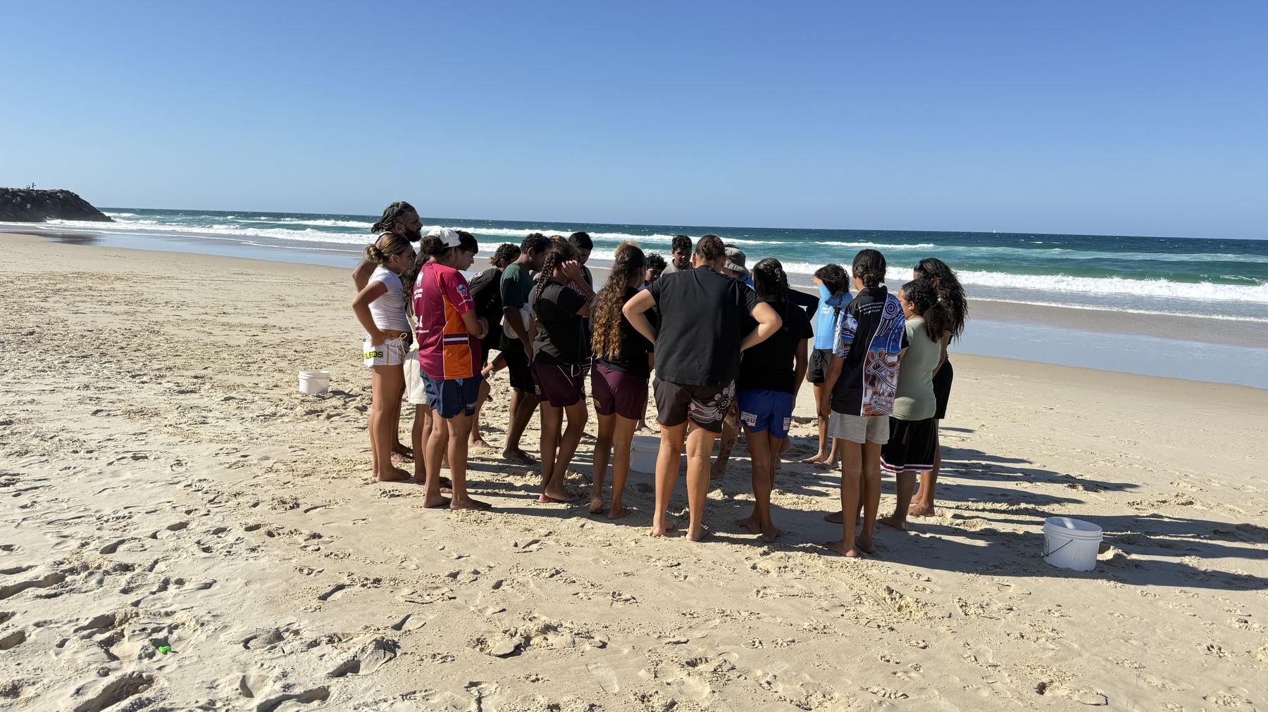 A circle of young people gathered on the beach for a yarn